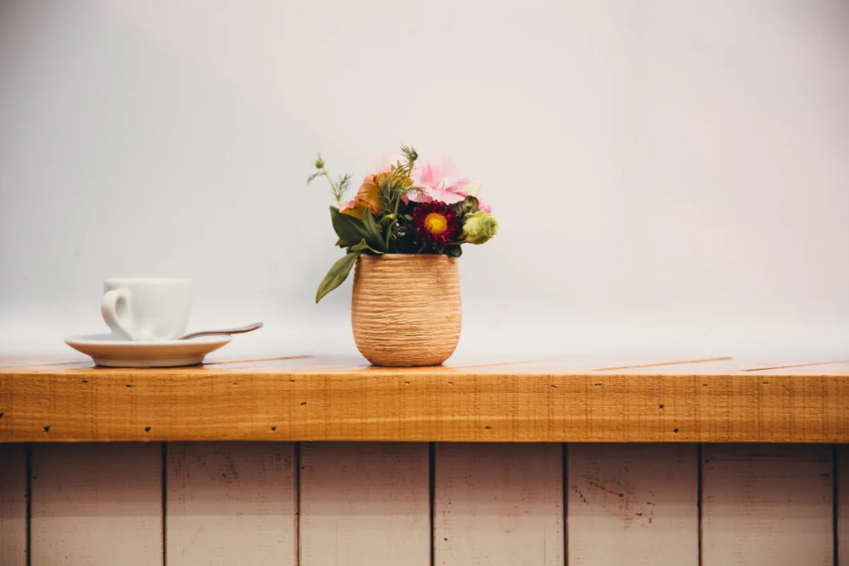 Una taza de café blanca sobre su plato y un pequeño jarrón de barro con flores de colores encima de una repisa de madera, frente a una pared clara