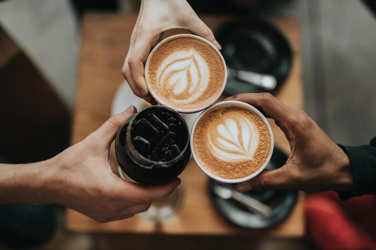 Tres manos acercando dos cafés con dibujo de hoja en la espuma y un vaso oscuro sobre una mesa de madera