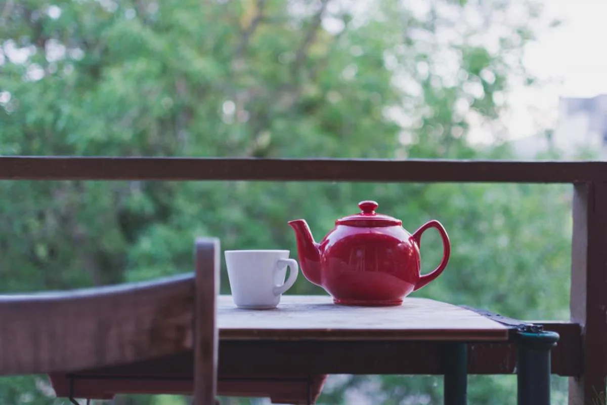 Tetera roja y taza blanca sobre una mesa de madera en un balcón con árboles verdes al fondo