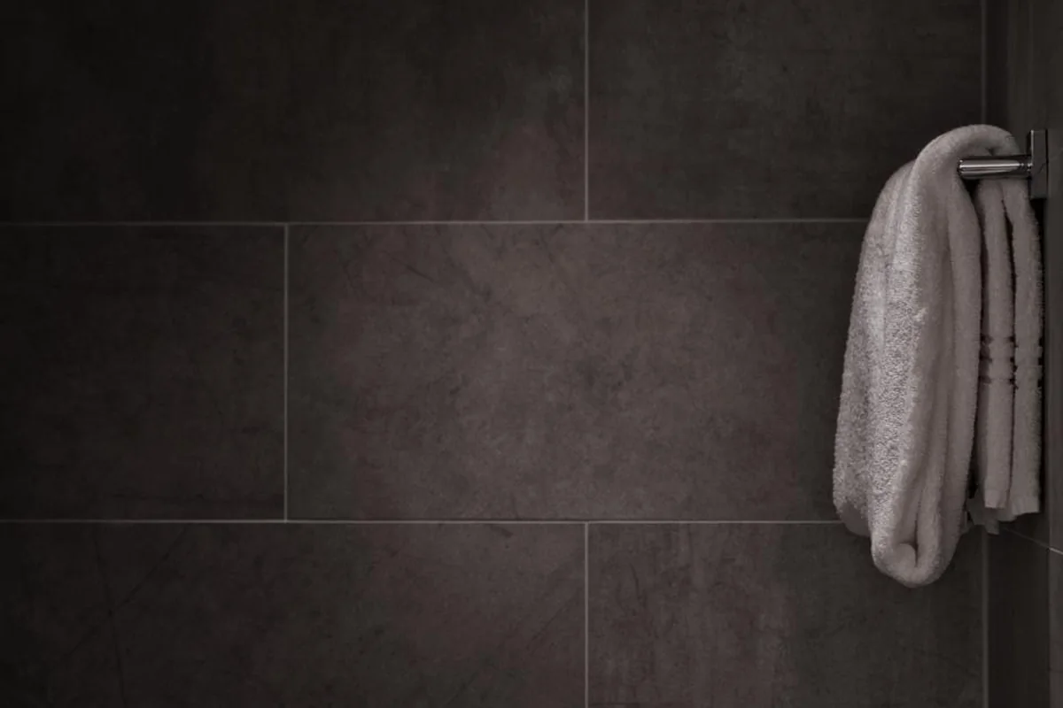 Toalla blanca doblada colgada de un toallero metálico en una pared de azulejos grandes de color gris oscuro