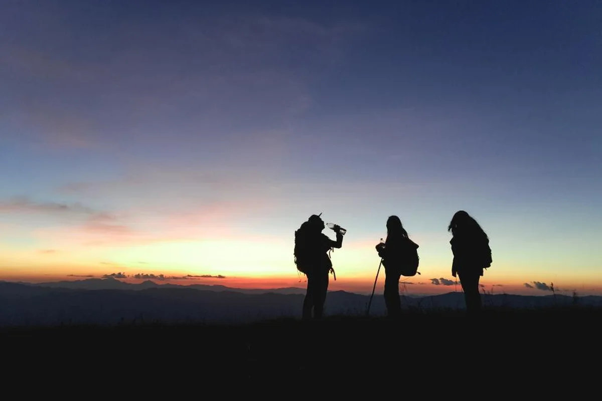 Tres siluetas de excursionistas con mochila en lo alto de una colina al atardecer