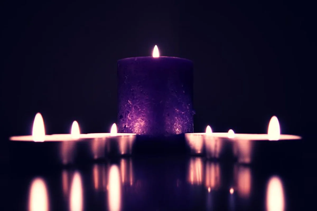 Vela gruesa de color morado encendida rodeada de pequeñas velas de té sobre una superficie que refleja su luz en la oscuridad