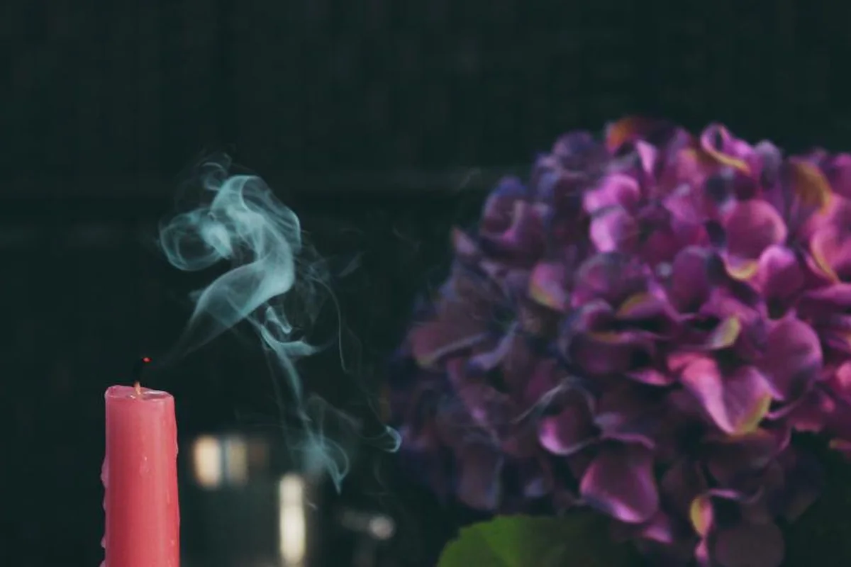 Una vela rosa recién apagada desprendiendo un hilo de humo junto a un ramo de hortensias moradas sobre fondo oscuro