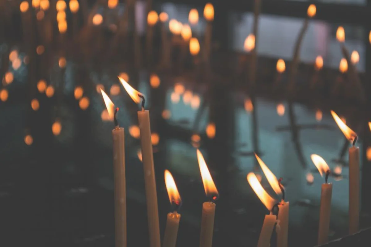 Varias velas finas y largas encendidas en primer plano, con muchas otras llamas desenfocadas al fondo en un espacio en penumbra