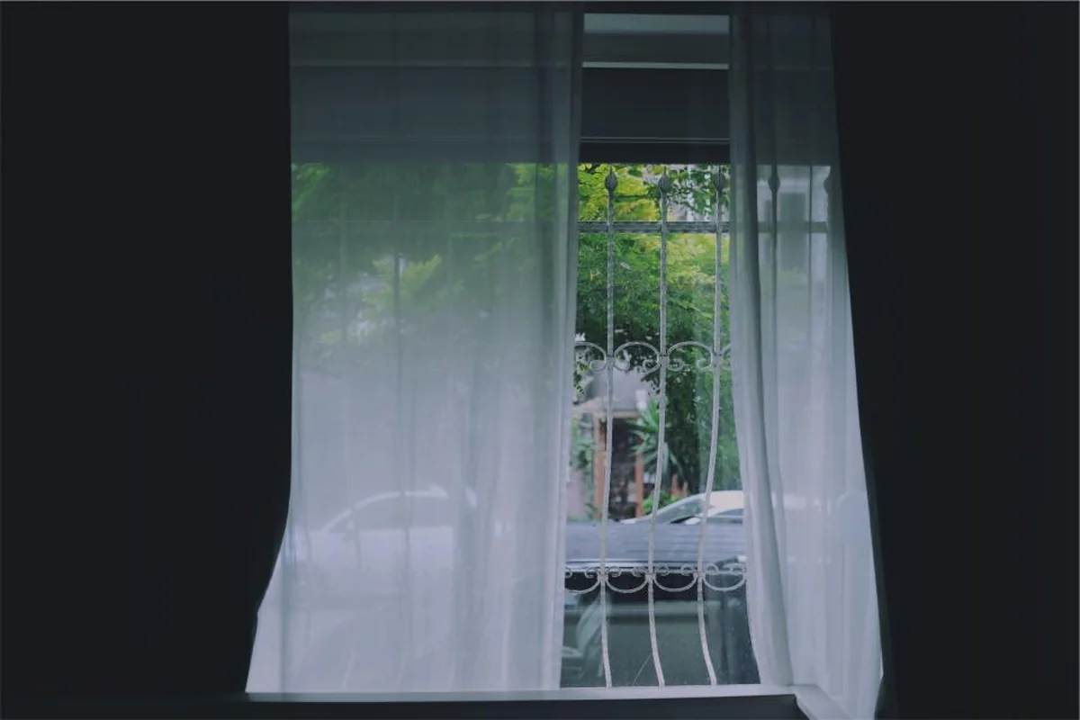 Ventana abierta con una cortina blanca y fina movida por el aire, con árboles y una reja de hierro al fondo