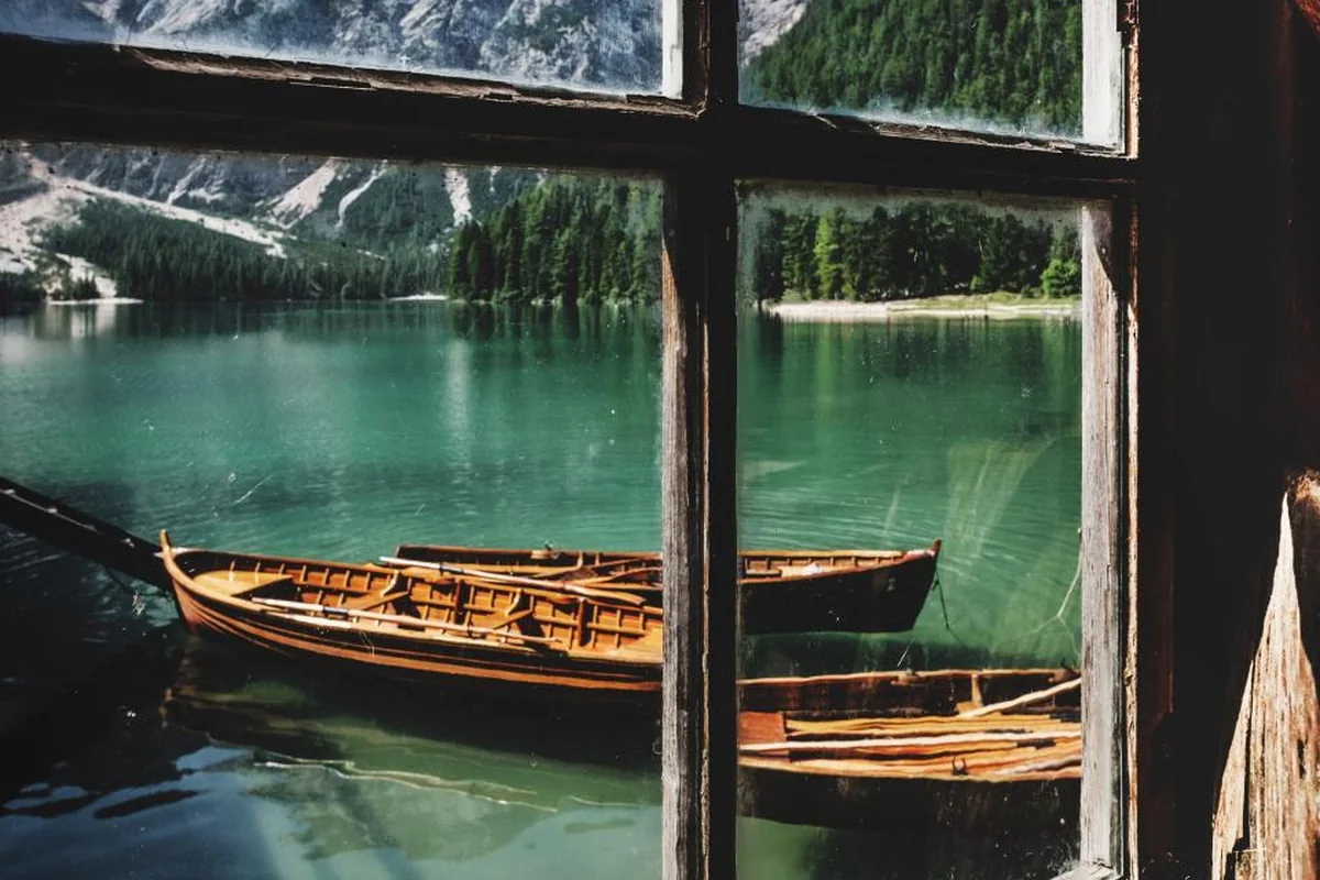 Barcas de madera sobre un lago de aguas verdes vistas a través de una ventana antigua de madera, con montañas al fondo