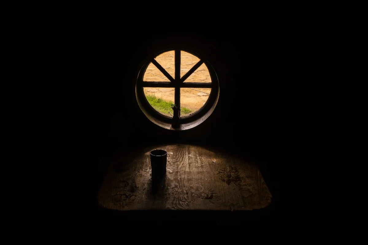 Ventana redonda iluminada por la luz del exterior en una habitación a oscuras con una mesa de madera y un vaso