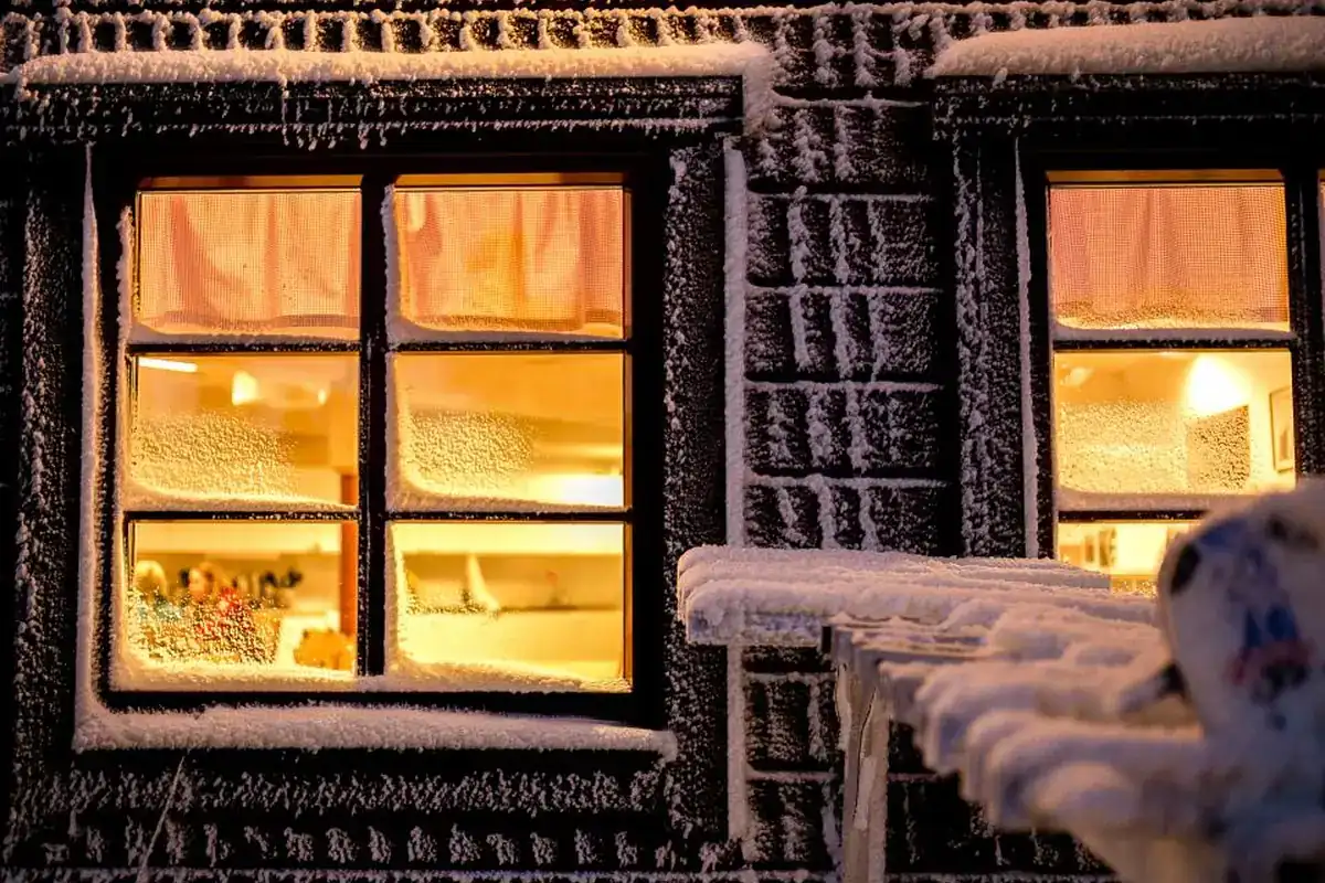 Dos ventanas con luz cálida en la fachada oscura de una casa cubierta de escarcha y nieve, de noche