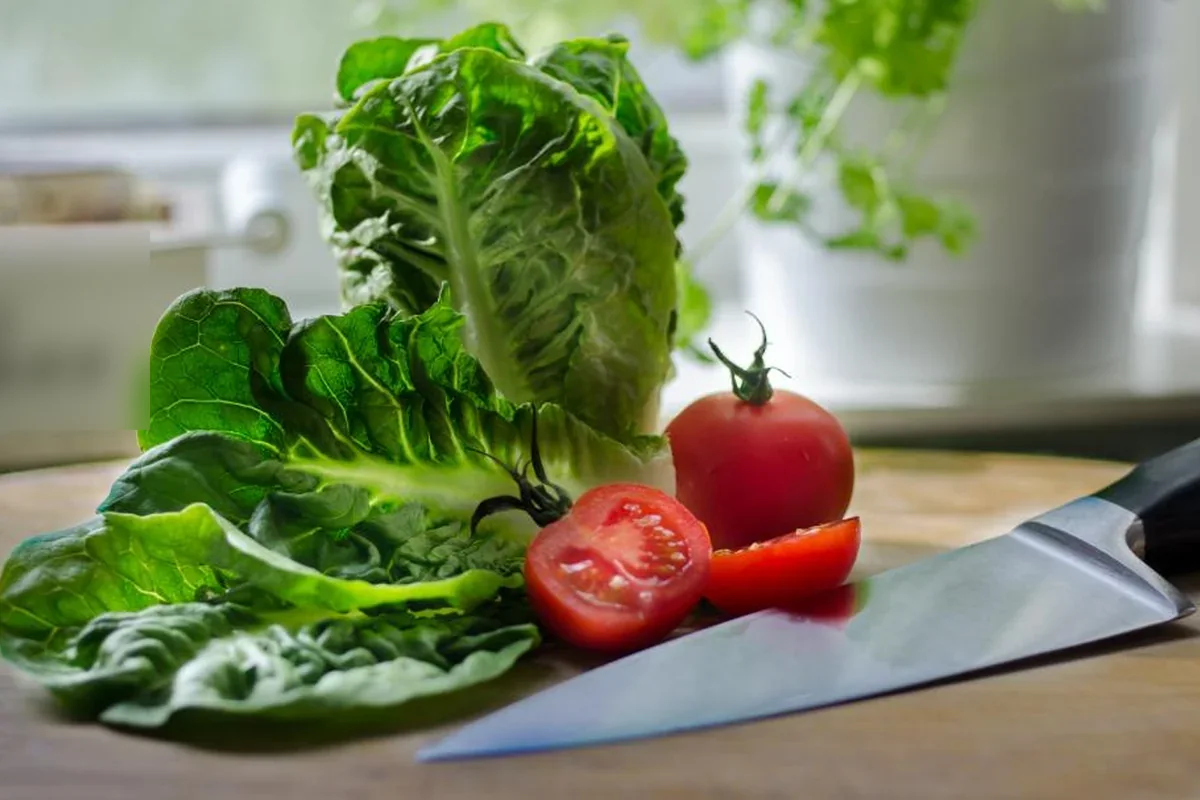 Hojas de lechuga romana, un tomate entero y otro cortado por la mitad junto a un cuchillo de cocina sobre una tabla de madera, con plantas desenfocadas junto a la ventana