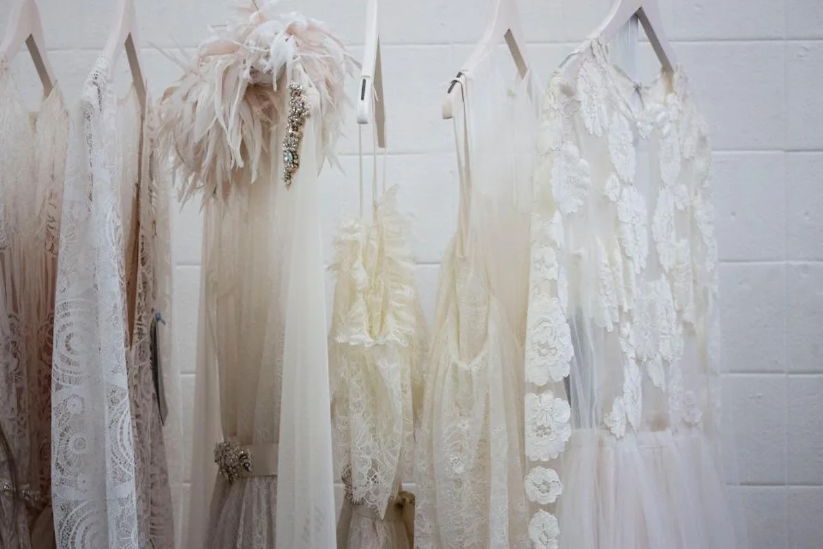 Varios vestidos y blusas blancas de encaje y tul colgados en perchas contra una pared de ladrillo pintada de blanco