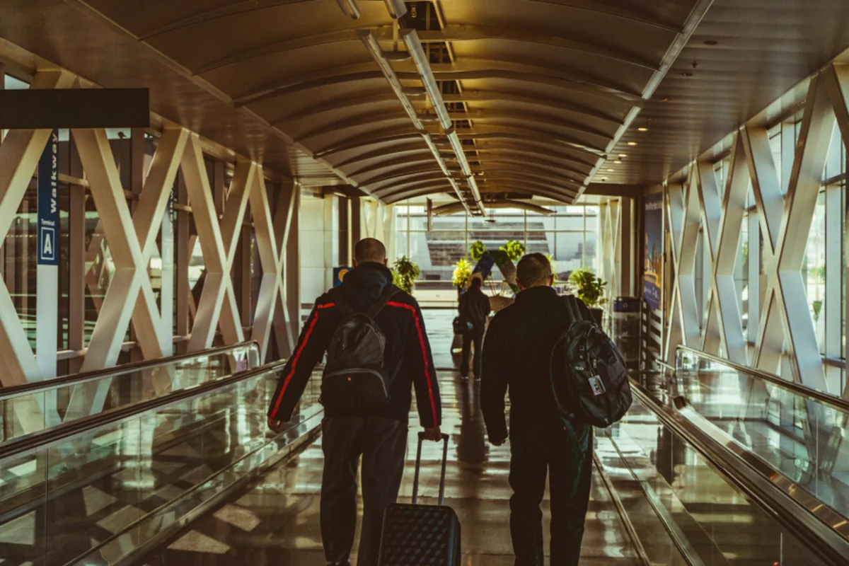 Dos personas de espaldas con mochilas y una maleta de ruedas caminando por una pasarela acristalada de aeropuerto