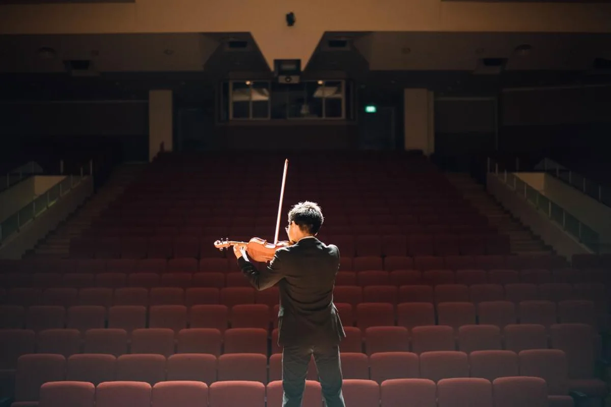 Violinista de espaldas tocando sobre el escenario frente a un auditorio con butacas rojas vacías