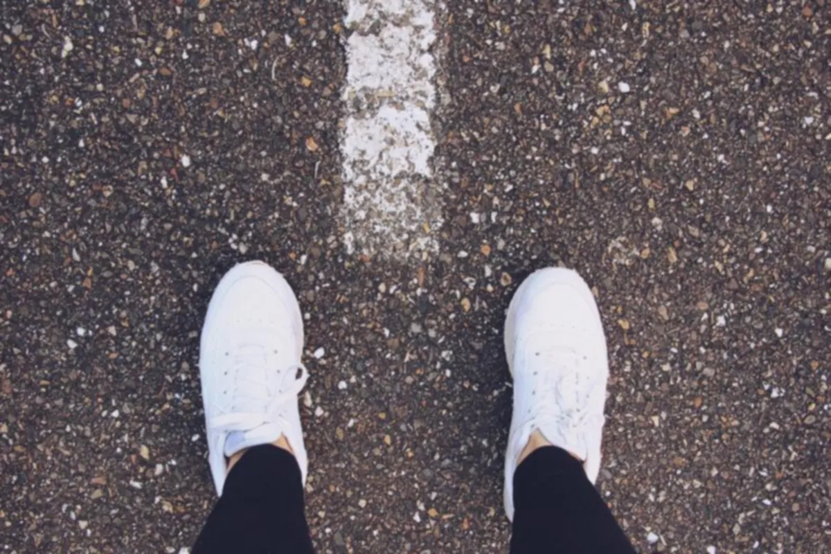 Pies con zapatillas blancas y pantalón negro sobre el asfalto, vistos desde arriba junto a una línea pintada