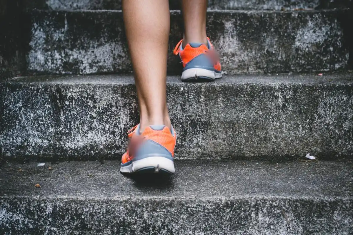 Piernas de una persona con zapatillas deportivas naranjas subiendo unos escalones de piedra gris